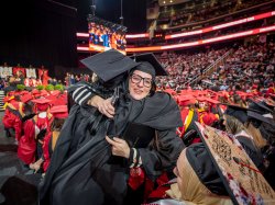 Two undergraduate students in black gowns and caps share a celebratory hug in the middle of the commencement floor.