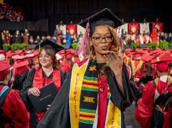 A Bloomfield College of Montclair State University graduate in a black gown and cap, wearing a colorful stole, walks down the aisle blowing a kiss amid rows of fellow graduates in red and black gowns.