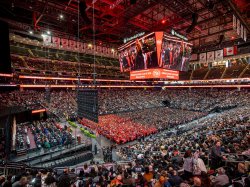 A wide view of the Prudential Center in Newark shows the arena filled with guests and rows of graduates in red and black gowns on the floor, as the commencement ceremony unfolds beneath large video screens hanging from the ceiling.