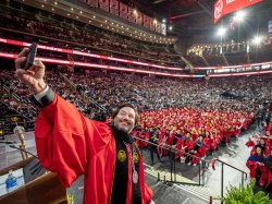 Montclair President Jonathan Koppell, wearing academic regalia, stands at the podium and raises his cellphone to take a selfie with a sea of graduates filling the arena behind him.