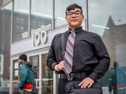Jose Carlos Aguilar, dressed in a black shirt and patterned tie, stands confidently outside a glass-fronted office building, holding a shoulder bag and phone, with blurred pedestrians and Paterson’s urban architecture in the background.