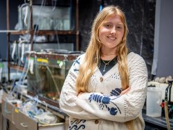 Kelly Witters stands in a marine biology lab with arms crossed, in front of stacked aquariums and tubing used for aquatic research.
