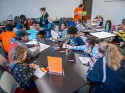 A classroom full of children and college students sit around large tables covered with markers, papers, and art supplies, as kids draw and craft while facilitators circulate and assist with the activities.