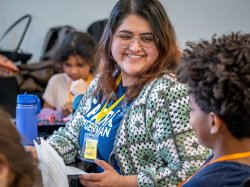 Isabella Zarate Gonzalez, wearing a blue SPARK T-shirt and a green-and-white crochet cardigan, sits at a table holding a worksheet, speaking with an elementary student who is seated around her during a classroom activity.