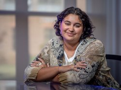 Dani Mazariegos sits at a studio desk with her arms folded, wearing a patterned blouse.