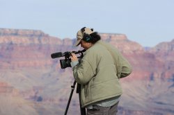 Dani Mazariegos stands outdoors on rocky terrain, wearing a light jacket and headset, as she adjusts a video camera on a tripod with desert cliffs and canyon walls in the background.