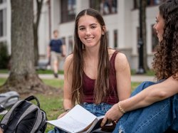 Dana Brzezinski sits on the grass at Montclair State University, holding an open notebook and talking with a friend, with campus buildings and a tree visible in the background.