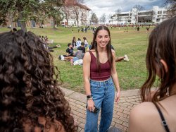 Dana Brzezinski stands on a grassy quad at Montclair State University, talking with two friends in the foreground while other students sit and gather in small groups across the lawn.