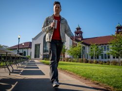 Casey Coleman walks along a campus pathway, holding a travel coffee mug, with long rows of empty folding tables on one side.