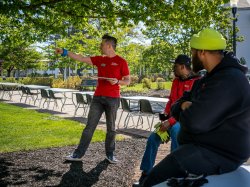 On a sunny morning under leafy green trees, Casey Coleman stands holding a clipboard and points across campus while two Facilities staff members sit on the edge of a table listening.