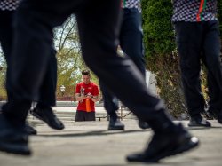 Casey Coleman stands in the background checking a phone while dancers in black pants and patterned tops move in a circle, their blurred legs framing the scene.