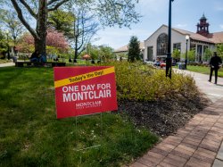 A red sign with while lettering says: One Day for Montclair. The campus is in background.