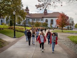 A campus tour guide and prospective students walk along a wide concrete path lined with grass, trees and lampposts on Montclair State University’s main quad, with a large white academic building with red tile roof and towers in the background.