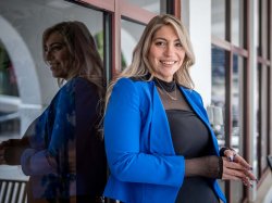 Ashley Crawford, in a black dress with bright blue blazer, stands in front of a building’s windows.
