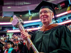 Grand Marshal Eric Diamond, a Montclair State University Theatre and Dance professor, wears academic regalia and holds the ornate silver University mace while leading the 2026 Spring Commencement procession inside Prudential Center.