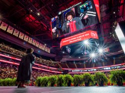 Montclair State University student Arianna Amina Joseph stands on the arena stage singing the national anthem into a microphone as the audience rises, while her image appears on the large Prudential Center video board above a banner reading “Congratulations Class of 2026.”