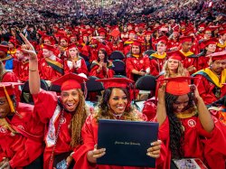 A large group of Montclair State University Graduate School students in bright red caps and gowns smile, cheer and hold up diplomas while seated together on the arena floor during the 2026 Spring Commencement ceremony, with rows of fellow graduates and guests filling the stands behind them.