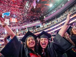 Two Montclair State University graduates in caps and gowns smile and raise their arms as red and white confetti rains down around them inside Prudential Center during the 2026 Spring Commencement ceremony.