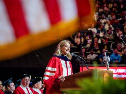 New Jersey Governor Mikie Sherrill, wearing red and white academic regalia, speaks at a podium during Montclair State University’s 2026 Spring Commencement, with a large American flag blurred in the foreground.