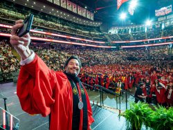 President Jonathan Koppell in red academic regalia holds up a phone to take a selfie with rows of graduating students in red gowns and a full arena of guests behind them during Montclair State University’s 2026 Spring Commencement.