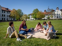 Students sit together on a blanket on a large grassy lawn at Montclair State University, talking and studying with notebooks and a laptop open, while academic buildings with red-tiled roofs and a clear blue sky frame the background.