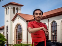 Transfer student John Bautista, a rising senior and Advertising major, sits confidently on a bright red chair in front of a Montclair State University building with white walls and red-tiled roofs, looking directly at the camera on a clear day.