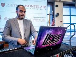 Ashon Lanada, wearing a gray blazer, sets up a laptop and sound equipment on stage, with a screen behind him displaying Montclair State University and partner college logos.