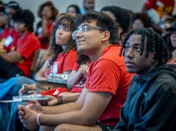Franco Leon, wearing a red EOF T-shirt sits with hands clasped, smiling and looking up attentively during a conference session.