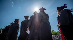 photo of graduates standing in black cap and gowns with blue sky and sunshine in background