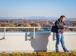 A student standing behind the CELS building reading a book with New York City in the distance.