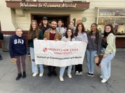 Students standing in front of a Farmers Market building holding a white Montclair State flag with red lettering