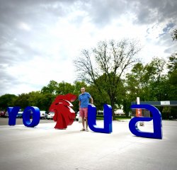 A man standing in front of giant metal letters that represent a University's name and logo.