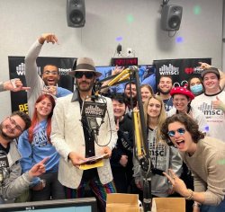A man with a beard wearing sunglasses and tophat surrounded by a group of smiling and excited students at a college radio station.