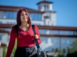 Oscaterin Bautista stands outdoors on the Montclair State University campus wearing a red long-sleeve shirt and black pants, carrying a shoulder bag. A large academic building visible in the background under a clear blue sky.