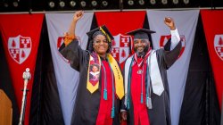 Father and daughter graduates, Lavone and Tiiera Broxton stand together with arms raised in their regaliaon Commencement stage with university banners behind them