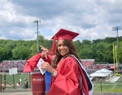 Josmary Medina smiling and waving to camera on graduation day in a red cap and gown.