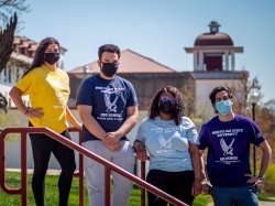 Four students standing near Red Hawk statue wearing masks and different colored t-shirts for different majors