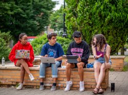 Four students sitting on brick ledge outside of Dickson Hall during the Summer Bridge Program researching different campus resources on laptops and notebooks.