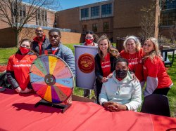 A photo of Montclair State students and faculty in red t-shirts at a table with a small spinning prize wheel