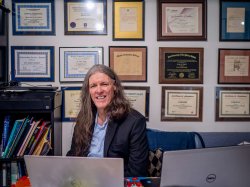 A man sits at his desk smiling with many diplomas on the wall behind him.