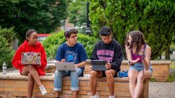 four students sitting on a low wall holding laptops and talking