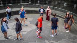 Group of nine students and a professor stand on a circular map of earth.