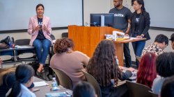 A woman in a pink blazer sits on a table speaking to a classroom of students.