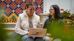 Two students are sitting in an outdoor courtyard, smiling and working on a laptop.