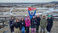 A group of students and professors stand with a Montclair State University pennant in front of a snowy landscape.
