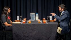 A man in a suit and tie gestures to a woman, both of whom are sitting in front of plates of chicken wings.