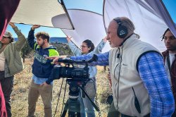 A professor standing by a video camera and wearing headphones instructs four students holding photo reflectors over their heads.