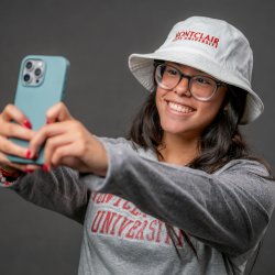 Woman student in glasses wearing a Montclair sweater and bucket hat combo while taking a selfie