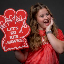 Woman student in red shirt holding a foam talon that reads Let's Go Red Hawks