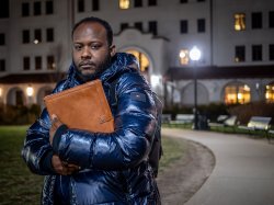 Horatio Wauchope,wearing a winter coat, stands outside on a campus walkway at night, holding a large folder to his chest in front of a lit academic building.
